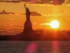 The sun rises with the Statue of Liberty in silhouette. Despite her troubles, there is a new dawn in American as President Biden restores environmental regulation.